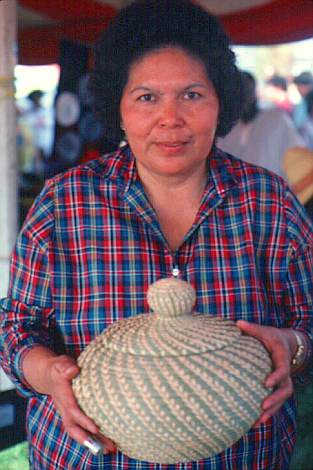 The Koasati Indians, also known as the Coushatta, of Elton have long produced coiled pinestraw baskets stitched with raffia and decorated with raffia and pinecones. While both the size of the coil and the spacing of the stitches vary, the closer the stitch and the smaller the coil, the stronger the basket is, and therefore, the higher quality the basket. Produced by many women in the tribe, these baskets have become an important source of community pride. Here, Marion John displays a pinestraw basket using the wheat stitch